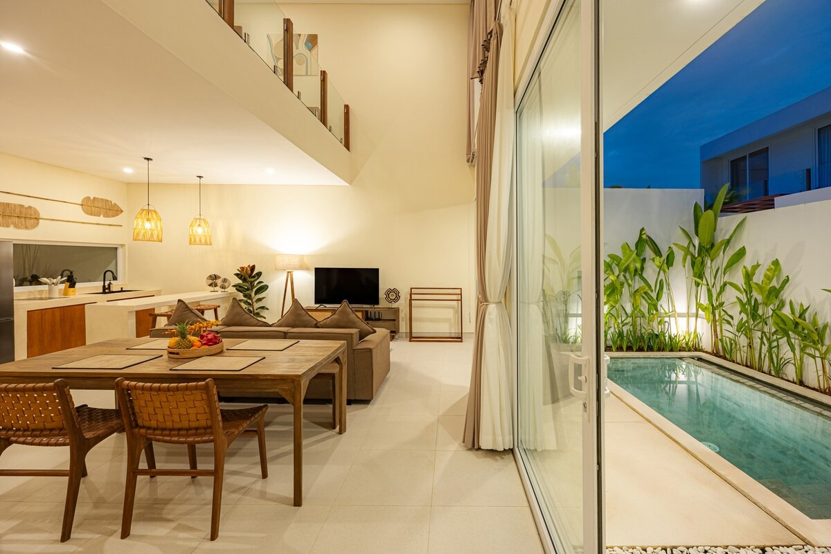 Open-plan living room with mezzanine above and the pool visible through the glass doors, at dusk